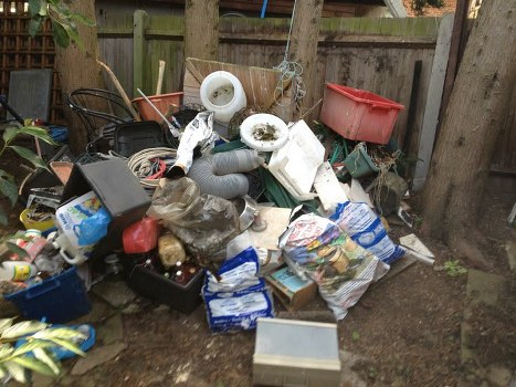 A large collection of assorted waste materials piled on a patch of bare ground within a backyard. The debris includes plastic bags, bags of rubbish, cardboard boxes, and various household items such as white plastic pipes and containers. Several black and red plastic bins and containers are visible among the clutter, with some partially hidden behind other objects. In the background, a weathered wooden fence and tree trunks frame the scene, indicating an outdoor residential setting. The waste appears unorganized and covers a significant area, suggesting the need for professional rubbish removal services like those provided by Commercial Waste Limehouse to clear such clutter efficiently. The scene is lit with natural daylight, highlighting the mixed textures and colors of the discarded items, emphasizing the typical waste accumulation encountered in domestic or garden cleanup situations in Limehouse or nearby areas.
