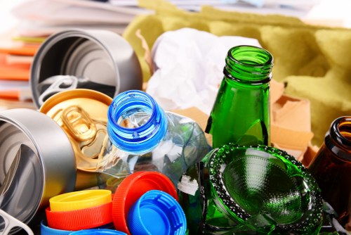 A close-up image shows a collection of various used glass and plastic bottles, bottle caps, and containers scattered on a surface, representing waste materials such as beverage bottles and lids. Among the objects, there are green glass bottles, clear plastic bottles with blue caps, and orange, yellow, and red bottle caps. The bottles are empty and some are partially crushed or crumpled. Visible in the background are other waste items, including paper and packaging materials, along with a partially visible cardboard box. The scene is set outdoors or in a waste collection environment, with natural or artificial lighting illuminating the scene. This image exemplifies typical waste materials that might be collected and handled by professional rubbish removal services, such as those provided by Commercial Waste Limehouse, to ensure proper disposal or recycling in Limehouse and surrounding areas post code E14.