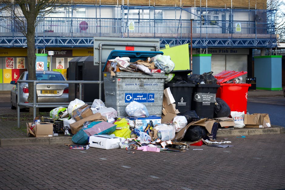 A waste collection truck with an open rear-loading compartment positioned on a narrow cobblestone street, surrounded by multi-story residential buildings with aged facades. The truck is primarily whit