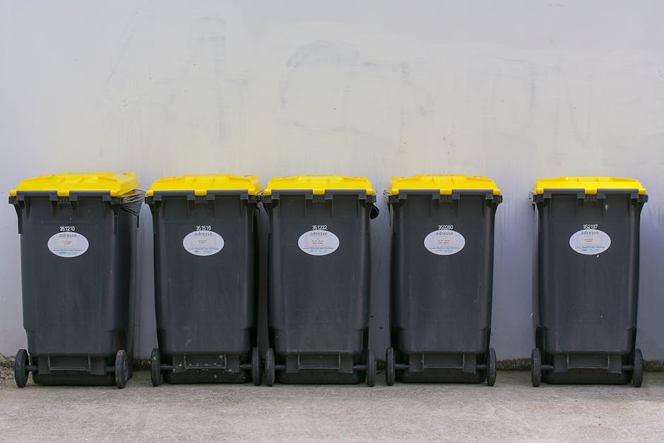 A man with dark hair, wearing a black T-shirt with white text, is shown leaning over and placing a white plastic bag filled with rubbish into a cylindrical outdoor waste bin made of shiny stainless st