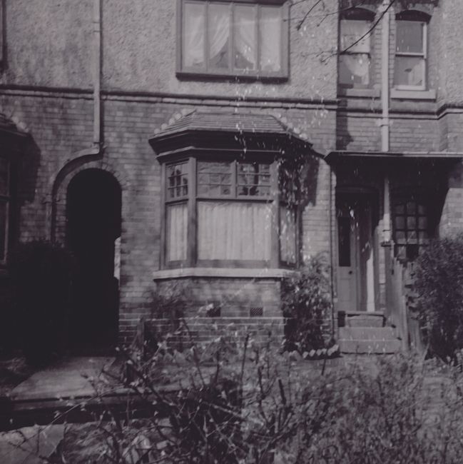 A black and white photograph of a two-storey brick terrace house with a protruding bay window on the ground floor, featuring wooden framing and multiple small panes of glass. The window has a slightly