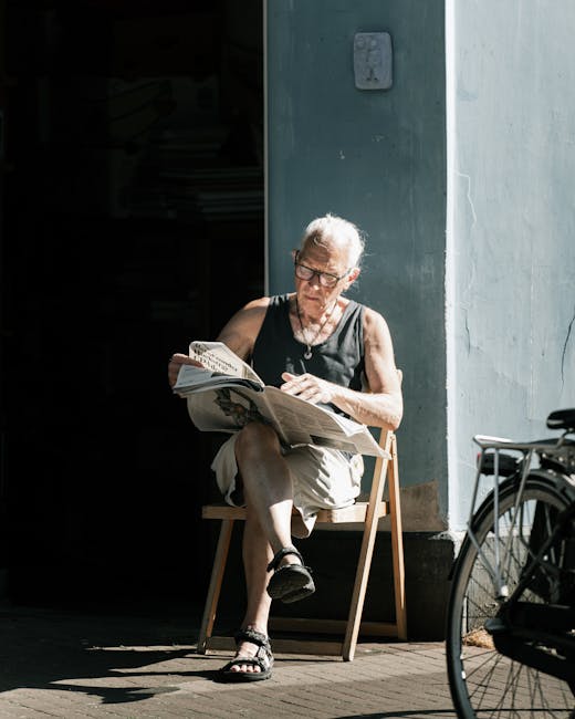 An elderly woman with short gray hair, wearing glasses, a sleeveless black top, and beige shorts, is seated outdoors on a small wooden chair next to a light blue wall with visible metallic utility box